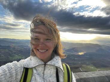 Una mujer sonriente con el pelo al viento está de pie en una montaña, vestida con un jersey grueso. El pintoresco fondo muestra colinas onduladas, valles y un espectacular cielo nublado.
