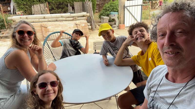 A group of six people, including adults and children, smile and pose around a white outdoor table. The background features yard equipment, wood, and greenery.
