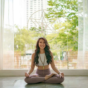 Uma jovem de cabelos longos está sorrindo enquanto está sentada de pernas cruzadas dentro de casa, praticando ioga ou meditação. A luz do sol entra pelas grandes janelas com árvores do lado de fora.