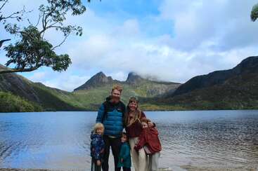 A happy family of five stands by a serene lake, surrounded by mountains and trees, under a partly cloudy sky, enjoying nature and each other’s company.