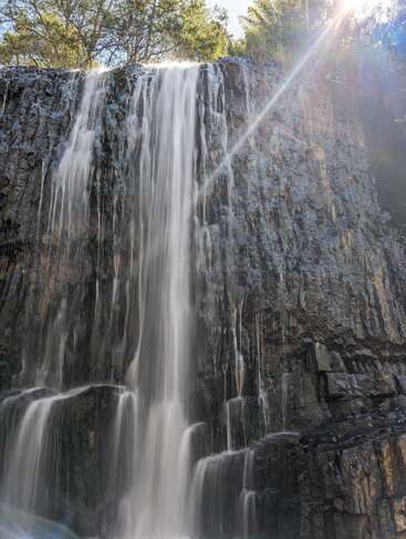 A majestic waterfall cascades down a rocky cliff, surrounded by lush greenery. Sunlight beams through trees, illuminating the mist, creating a serene and refreshing natural scene.