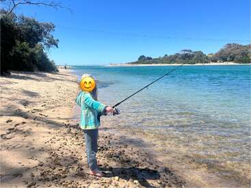A child stands barefoot on a pebbly beach, fishing by clear blue water under a sunny sky. Trees and distant shorelines create a peaceful, relaxing scene.