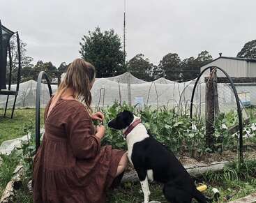 A woman in a brown dress sits in a garden, tending to plants. A black and white dog sits beside her, observing. Overcast sky above.