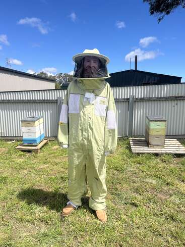 A person in a protective beekeeping suit stands on grass near several beehives, surrounded by metal fencing, under a bright blue sky with clouds.