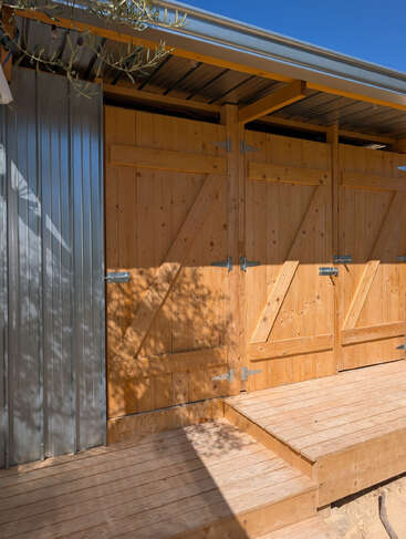 This image shows a wooden structure with double doors, a corrugated metal wall, and a wooden ramp. Sunlight casts shadows of tree branches across the surfaces.