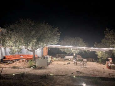 An outdoor seating area at night under trees with string lights, pallet sofas, tables, chairs, and a tarp canopy, creating a cozy, rustic atmosphere.