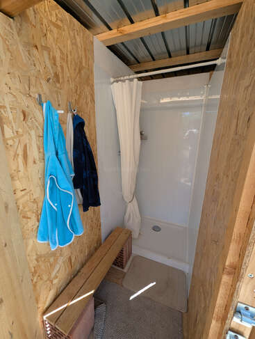 This image shows a small, rustic shower room with wooden walls, a simple curtain, a bench, two hanging robes, a mat, and a corrugated metal roof.