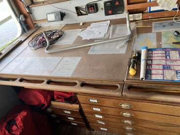 Ein hölzerner Navigationstisch mit Karten, Papierkram, Werkzeug und Kabeln. Darunter befinden sich mehrere Schubladen, in denen rote Jacken zu sehen sind. Der Arbeitsbereich wirkt organisiert und maritim.
