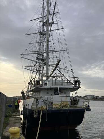 Ein großes Segelschiff liegt am Kai, seine Masten und Takelage ragen in den wolkenverhangenen Himmel. Menschen stehen auf dem Steg daneben.