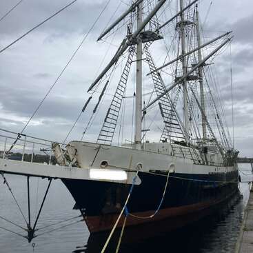 Ein großes, altes Segelschiff mit hohen Masten liegt an einer Anlegestelle. Der Himmel ist bedeckt und bietet eine dramatische Kulisse für das historische Schiff.