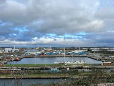 Das Bild zeigt einen Industriehafen unter einem wolkenverhangenen Himmel, mit Lagerhäusern, angedockten Booten und grünen Flecken. Das Wasser spiegelt die Szene und unterstreicht ihre ruhige Atmosphäre.