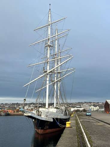 A tall ship with three masts is docked at a quiet harbor. The sky is overcast; buildings, a blue car, and industrial structures are visible nearby.