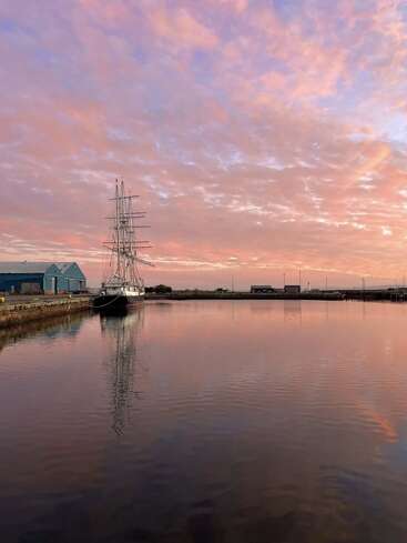 Un grand navire est amarré à côté d'entrepôts bleus, se reflétant sur une eau calme. Le ciel est rempli de nuages doux et colorés lors d'un coucher de soleil serein.