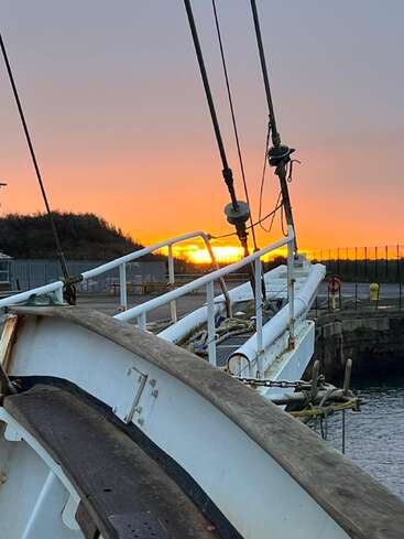 Un navire amarré au port est vu au coucher du soleil, avec des teintes orange et jaunes remplissant le ciel, jetant une lueur chaleureuse sur la scène.