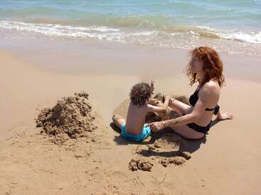 A woman and a child sit on the beach, building a sandcastle together, with the ocean waves gently lapping at the shore in the background.
