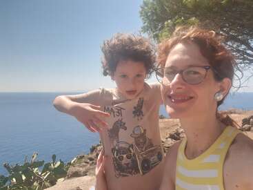 The image shows a woman and a child posing for a selfie on a cliff overlooking the ocean, with a tree and cactus plants in the foreground.