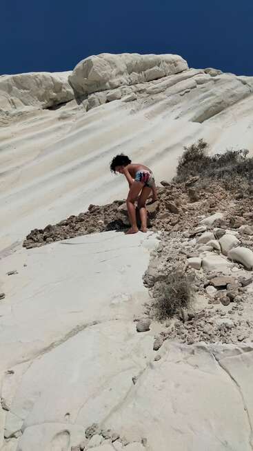 A child in swim trunks climbs up a steep, white rocky hill under a clear blue sky, surrounded by dry bushes and rugged, sunlit terrain.