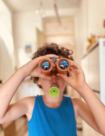 A young child with curly hair wears a blue tank top, looking through orange toy binoculars and blowing a green whistle indoors, appearing curious and playful.