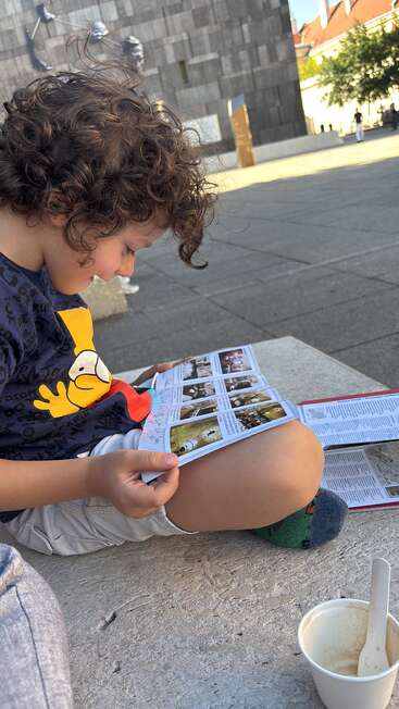 A young boy with curly hair sits outdoors on a stone bench, reading a magazine. An empty ice cream cup rests nearby, urban buildings in the background.