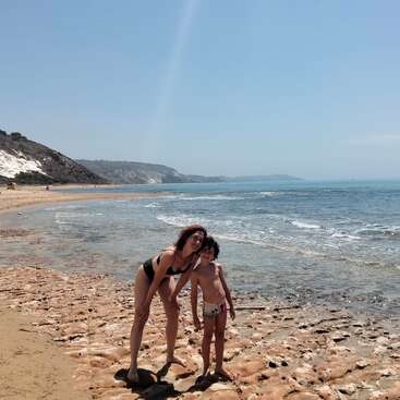 A woman and a child pose together on a sandy beach, with rocky shorelines, waves, white cliffs, and distant hills under a bright blue sky.
