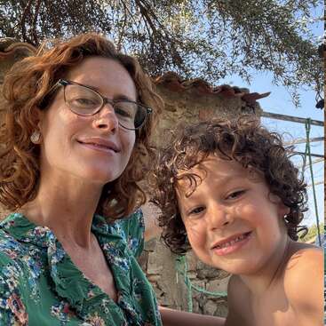 A woman with curly red hair and glasses smiles beside a young curly-haired boy. They appear outdoors in sunny weather, with stone walls and trees behind them.