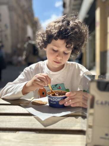 Un enfant aux cheveux bouclés est assis dehors à une table en bois, tout occupé à manger une glace au chocolat dans un pot bleu, le visage et son environnement baignés par la lumière du soleil.