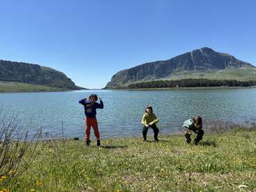 Trois enfants jouent au bord d'un lac paisible, entourés d'herbe verte et de fleurs sauvages. En arrière-plan, des montagnes majestueuses se dressent sous un ciel bleu limpide.