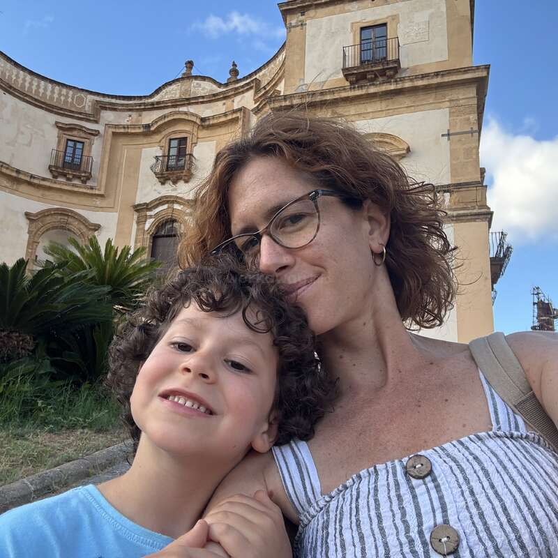 Une femme et un jeune garçon sourient ensemble pour un selfie devant un magnifique bâtiment ancien aux fenêtres cintrées, entouré de verdure, sous un ciel d'un bleu éclatant.