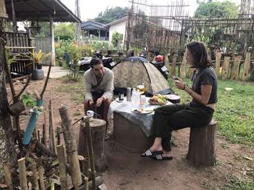 A imagem retrata um ambiente externo sereno, com um homem e uma mulher sentados em uma mesa com uma tenda ao fundo, cercados por uma vegetação exuberante e uma atmosfera rústica.