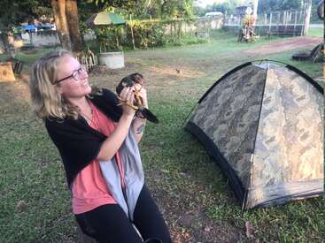 Uma mulher segura um pássaro em frente a uma barraca em uma área gramada, com árvores e outras estruturas ao fundo em um dia ensolarado.