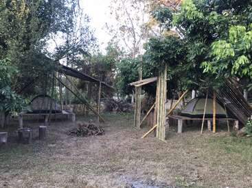 A imagem retrata um ambiente externo rústico com uma tenda, estruturas de madeira e uma área gramada, cercada por árvores e folhagens, evocando uma sensação de tranquilidade natural.