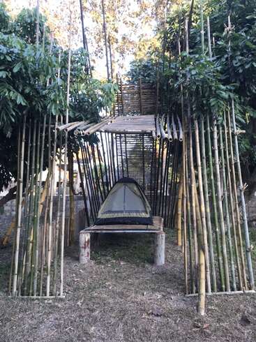 A imagem mostra um ambiente externo sereno com uma tenda aninhada em uma estrutura de bambu, cercada por uma vegetação exuberante e uma área gramada.
