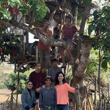 The image depicts a group of people posing for a photo in front of a treehouse, with some standing on the tree\'s branches.