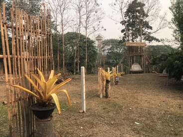 A imagem mostra um ambiente externo sereno com uma área gramada, cerca de bambu e vasos de plantas, com uma estrutura em construção ao fundo.