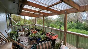 This covered wooden porch is filled with various potted plants, gardening tools, and crates. It overlooks a lush, green garden surrounded by trees and nature.