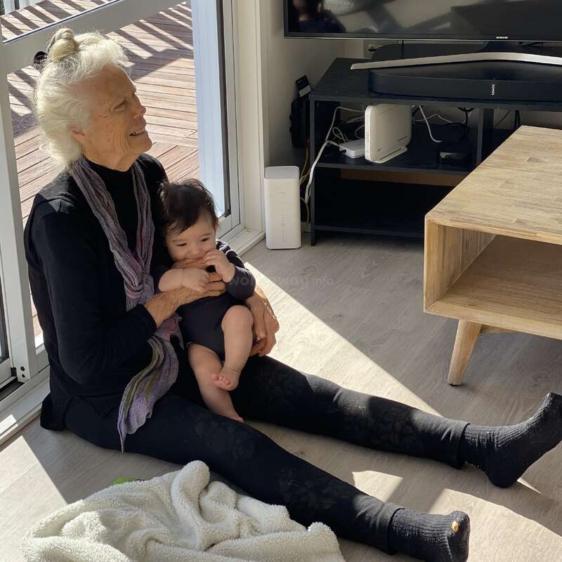An elderly woman sits on the floor, cradling a baby, with a TV and coffee table nearby, in a room with a deck outside.