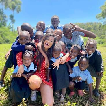 Das Bild zeigt eine Frau in einem roten Kleid, die mit einer Gruppe von Kindern auf einer Wiese kniet und an einem sonnigen Tag für ein Foto posiert.