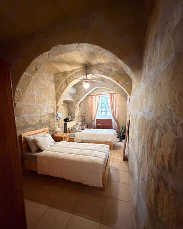 Dieses Bild zeigt ein gemütliches Schlafzimmer mit Steinwänden, zwei Einzelbetten, gewölbten Decken, Fliesenboden, sanfter Beleuchtung, einem Deckenventilator und einem Fenster mit rosa Vorhängen.