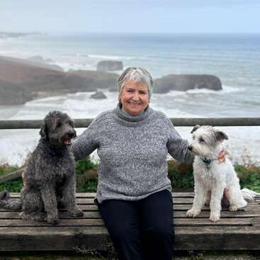 An older woman sits on a wooden bench between two dogs, smiling. Behind them, the ocean waves crash against rocky cliffs under a cloudy sky.