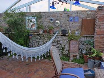 Une terrasse chaleureuse avec un hamac blanc, des plantes en pot, des murs rustiques en brique et en pierre, des coussins bleus, un miroir décoratif et des outils de jardinage, idéale pour se détendre en plein air.