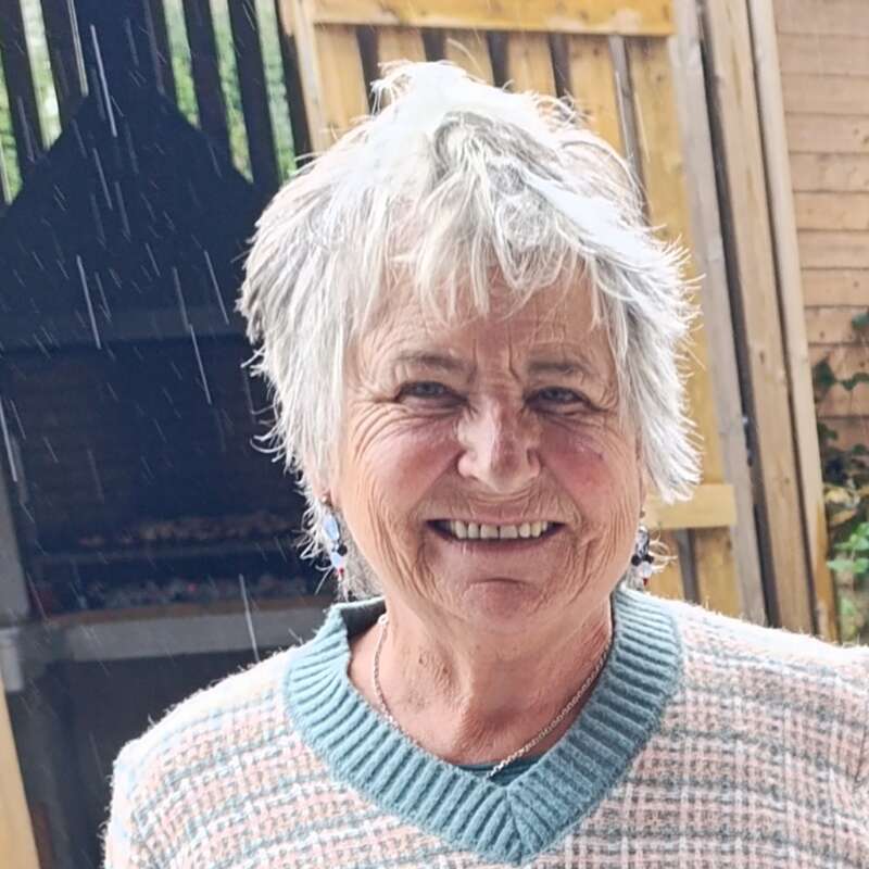 An elderly woman with short white hair smiles warmly. She wears a plaid sweater and earrings, standing outdoors in light rain, with wooden fences and greenery behind her.