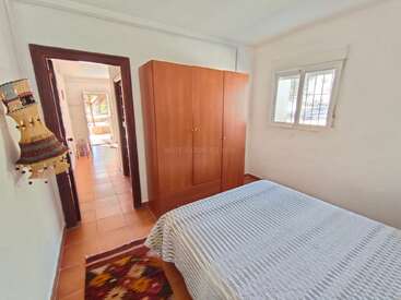 La chambre est simple et lumineuse, avec un lit recouvert d'une couverture blanche, une armoire en bois, un tapis coloré, une fenêtre, des décorations murales et un sol carrelé.