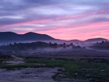 Un paysage serein au crépuscule, avec des collines ondulantes se découpant sur un ciel rose et violet, la brume recouvrant doucement le sol, créant une atmosphère calme et rêveuse.
