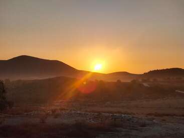 L'image montre un paysage serein au coucher du soleil, avec le soleil se couchant derrière des collines ondulantes. Une lumière dorée et chaude baigne le ciel et le terrain rocheux ouvert.