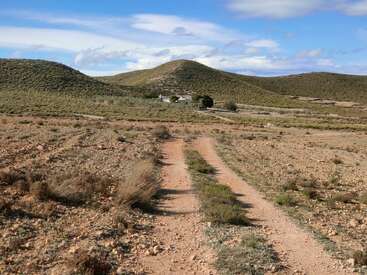 Un étroit chemin de terre serpente à travers un terrain aride et rocheux, menant vers de lointaines collines verdoyantes et une petite maison sous un ciel bleu parsemé de nuages.