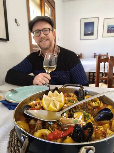 Un hombre con gafas y gorra sonríe mientras sostiene una copa de vino blanco. Delante, en un restaurante, se sirve una apetitosa paella de marisco con limones.