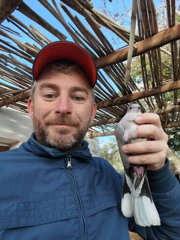 Un hombre vestido con una chaqueta azul y una gorra roja sostiene un pájaro blanco y gris en su mano derecha, sonriendo, bajo una rústica pérgola de madera al aire libre.