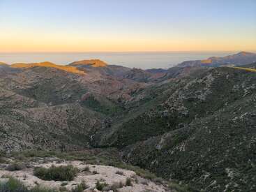 Paisaje montañoso bajo un cielo despejado, iluminado por la luz dorada del sol. Las colinas onduladas descienden hacia verdes valles, y el horizonte lejano se encuentra con un mar tranquilo y sereno. Atmósfera pacífica y serena.