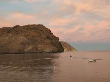 Una tranquila escena al atardecer muestra un escarpado acantilado costero, las tranquilas aguas del mar, una pequeña embarcación y una moto acuática, bajo un cielo de tonos pastel con suaves nubes.