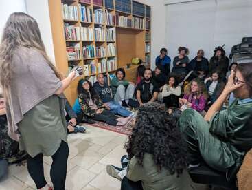 A group of people sits closely together on the floor and chairs, attentively listening to a woman speaking in a cozy room filled with bookshelves.